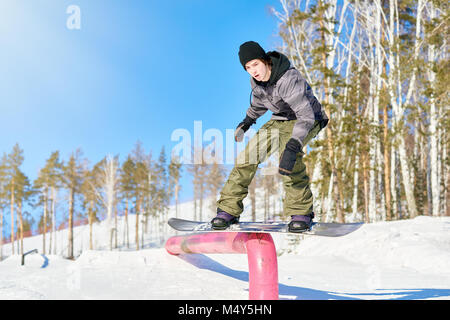 Volle Länge Action Shot des jungen Mannes, Durchführung von Snowboard stunt Hineinrutschen Metallgeländer im Sonnenlicht am Ski Resort, kopieren Raum Stockfoto