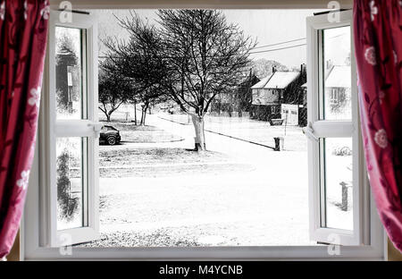 Blick durch die offenen Fenster auf einen schönen Winter Schnee Straße Szene im ländlichen England. Rote Vorhänge hängen vor dem modernen Doppelt verglaste Fenster Stockfoto