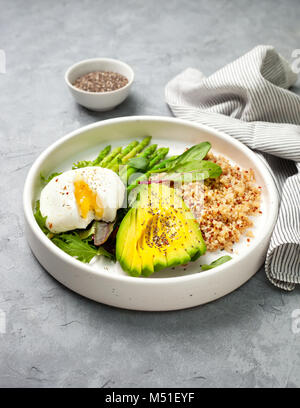 Gesunde Ernährung Frühstück. Quinoa, Avocado, Spargel, pochierte Eier in einem weißen Keramikplatte auf grauem Stein Hintergrund Stockfoto