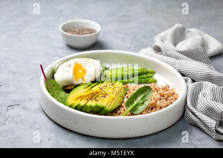 Gesunde Ernährung Frühstück. Quinoa, Avocado, Spargel, pochierte Eier in einem weißen Keramikplatte auf grauem Stein Hintergrund Stockfoto