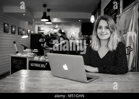 Ein junger Junge Frau schreibt ein blog aus einem Café in eine alternative und moderne Art zu Arbeiten auf Ihrem Laptop beim Trinken einen Kaffee Stockfoto