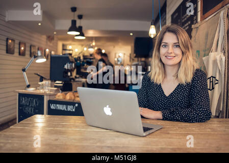 Ein junger Junge Frau schreibt ein blog aus einem Café in eine alternative und moderne Art zu Arbeiten auf Ihrem Laptop beim Trinken einen Kaffee Stockfoto