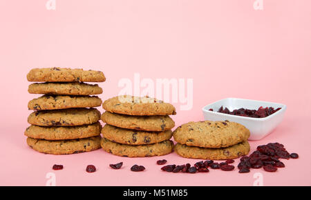 Stapel von oatmeal cranberry Cookies auf ein rosa Oberfläche und Hintergrund. Eckige Schale mit getrockneten Cranberries hinter Stacks mit zufälligen Preiselbeeren auf t Stockfoto