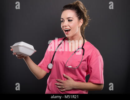 Portrait von schönen jungen Arzt holding Lunch Box suchen glücklich auf schwarzem Hintergrund Stockfoto