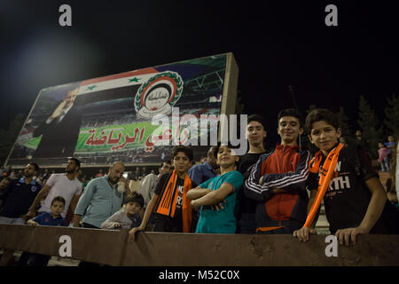 Jungen posieren vor einer Plakatwand mit dem syrischen Präsidenten Baschar al-Assad in der Syrischen Fußball-Cup Finale in Tishreen Stadion. Die syrischen Fußball WM-Finale wurde zwischen Al-Wahda und Al-Karamah Fußball-Mannschaften gespielt. Trotz des anhaltenden Konflikts in der vom Krieg zerrissenen Land, in Syrien, dem syrischen Fußball Cup fand in der Hauptstadt Damaskus, die unter der syrischen Regierung Steuerung ist mit vielen einheimischen Fußball-Fans das Match besucht. Stockfoto