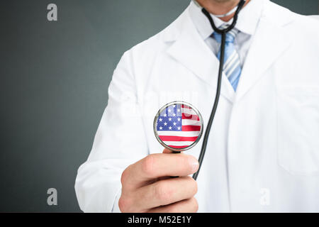 Mittelteil der männlichen Arzt Holding Stethoskop mit amerikanischer Flagge auf Chestpiece gegen grauer Hintergrund Stockfoto