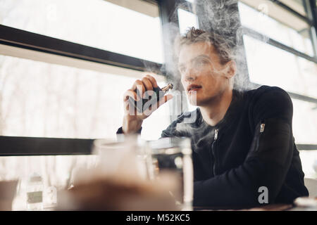 Junger Mann vaping in geschlossenen öffentlichen Raum. Rauchen elektronische Zigarette im Cafe. Nikotinsucht. Weise zu rauchen zu beendigen, alte Gewohnheit. Vaping Aroma, urbane Menschen u Stockfoto