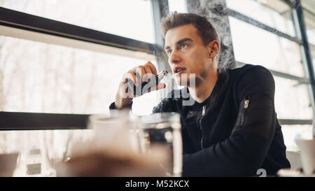 Junger Mann vaping in geschlossenen öffentlichen Raum. Rauchen elektronische Zigarette im Cafe. Nikotinsucht. Weise zu rauchen zu beendigen, alte Gewohnheit. Vaping Aroma, urbane Menschen u Stockfoto