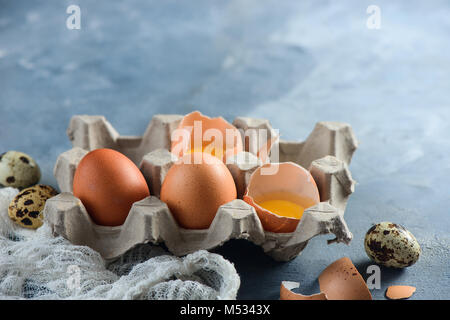 Frische Wachteln und Huhn Eier, ganze und gebrochen, in einem Papier Verpackung auf einen konkreten Hintergrund. Zutaten für Ostern kochen. Close-up mit kopieren Stockfoto