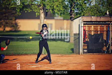 Ein Teenaged stehendes Mädchen zu Hause Platte in einem schwarzen Baseball uniform und Helm Schwingen einer bat auf einen eingehenden gelb Baseball Stockfoto