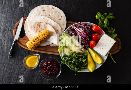 Tortillas flach und verschiedene Gemüse für Tacos oder Burrito auf Holzbrett auf schwarzem Schiefer Tabelle, Ansicht von oben, Hintergrund