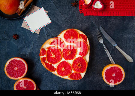 Ansicht von oben gebackene Käsetorte mit roten Pampelmuse Scheiben in der Form von Herzen auf den schwarzen Stein, Postkarte für Wünsche, frische Früchte und Besteck. Selektiver Fokus, Platz für Text. Stockfoto