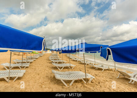 Weißen Chaiselounges und blaue Schirme stehen am Strand im Sand in der Nähe des Ozeans mit grünen Wellen und ein blauer Himmel mit weißen Wolken Stockfoto