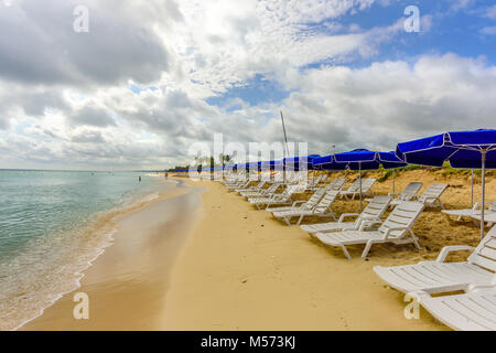 Weißen Chaiselounges und blaue Schirme stehen am Strand im Sand in der Nähe des Ozeans mit grünen Wellen und ein blauer Himmel mit weißen Wolken Stockfoto