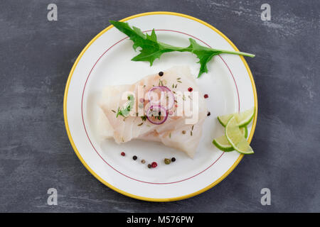 Frische rohe cod Fischfilet auf einer Platte mit Petersilie und Zitrone auf grauem Schiefer Hintergrund isoliert Stockfoto