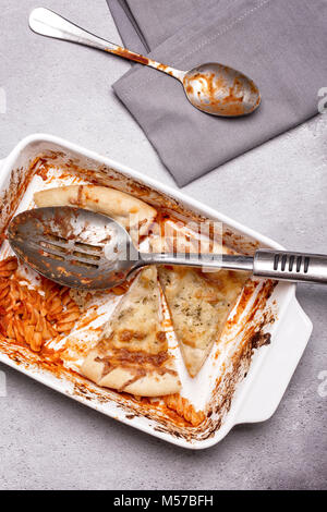 Teller der übrig gebliebenen Nudeln backen und kitschiger Knoblauchbrot mit einem Löffel und Servietten auf grauem Hintergrund im Landhausstil Stockfoto