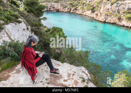 Eine ruhige warten Junge brünette Frau mit ergrauendem Haar, einen roten Pullover und Schal, der auf einem Felsen an der Steilküste der Cala Pi sitzen auf der Insel Mallorca Stockfoto