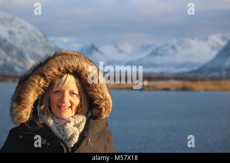 Nahaufnahme der blonde Frau mittleren Alters tragen Snood, grau Parka mit Fell Kapuze mit Schnee Berge und Meer im Hintergrund in der Sonne abgedeckt Stockfoto