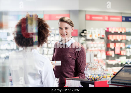 Porträt von einem gutaussehenden jungen Mann lächelnd beim Kauf eine nützliche Arzneimittel in einer modernen Apotheke mit verschiedenen Arzneimitteln und hilfsbereite Person Stockfoto