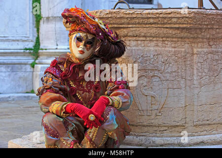 Ein Harlekin neben einem alten Brunnen während des Karnevals von Venedig (Carnevale di Venezia) in Venedig, Italien sitzt Stockfoto