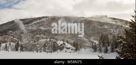 Panoramablick auf die Landschaft der schneebedeckten Feld gegen Roaring Mountain mit aktiven geysir Dampföffnungen mit Pinien gegen den blauen Himmel im Yellowstone Na Stockfoto