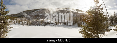 Panoramablick auf die Landschaft der schneebedeckten Feld gegen Roaring Mountain mit aktiven geysir Dampföffnungen mit Pinien gegen den blauen Himmel im Yellowstone Na Stockfoto