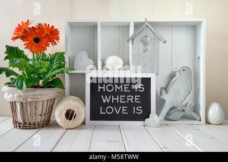 Frühling Hintergrund mit Frühling Dekorationen. Vitrine mit verschiedenen Objekten und Orange Gerbera Flower Pot. Text 'Heimat ist, wo Liebe' auf einem ist Stockfoto