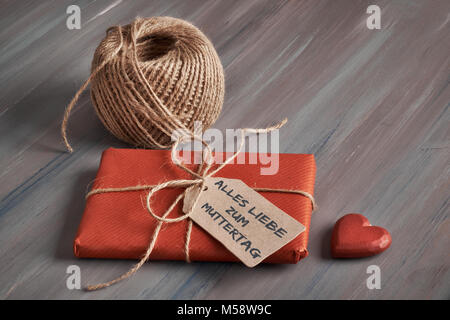 Verpackte Geschenk mit Schnur, Karton tag mit Text "Alles Liebe zum Muttertag" und eine hölzerne Herz auf rustikalen Holztisch. Der Text bedeutet "Alle Stockfoto