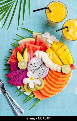 Tropische Früchte-Sortiment auf einem weißen Teller mit Palmblatt-Baum mit Mango-Smoothie, Textile blauen Hintergrund. Ansicht von oben. Textfreiraum Stockfoto