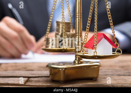 Nahaufnahme des Modell home und Münzen auf goldenen Waage mit Geschäftsmann im Hintergrund Stockfoto