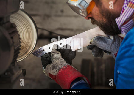 Ein Handwerker in der Werkstatt eine Neu geschliffenen Stück Stahl mit dicken Handschuhen und Glättung und Veredelung das Ende mit einer Fläche Schleifer. Tragen Stockfoto