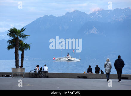 Die Menschen am Ufer am Genfer See in Lausanne mit Bootsfahrt und die Berge im Hintergrund. Stockfoto