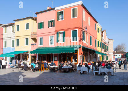 Insel Burano, Venedig, Venetien, Italien. Menschen speisen in einem Open-air-restaurant in der Via Baldassare Galuppi im Winter mit hellen bunten Gebäude Stockfoto