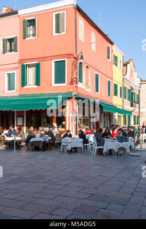 Insel Burano, Venedig, Venetien, Italien. Diners in der Via Baldassare Galuppi sitzen an einem Open air Restaurant in der Wintersonne Stockfoto