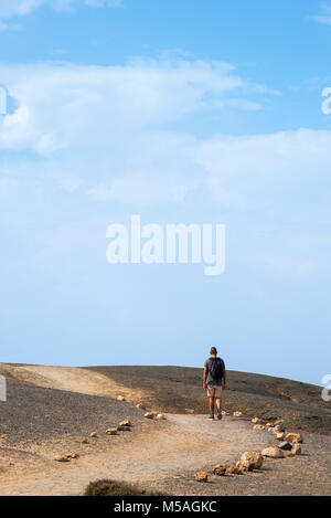 Ein junger weißer Mann, von hinten gesehen, mit einem Rucksack zu Fuß durch eine vulkanische Landschaft in Fuerteventura, Kanarische Inseln, Spanien Stockfoto