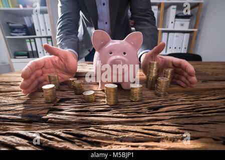 Nahaufnahme der Kaufmann schützende Hand gestapelte Münzen und Sparschwein auf hölzernen Schreibtisch Stockfoto