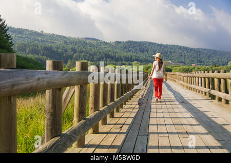 Junge Frau zu Fuß auf die hölzerne Weg entlang dem Seeufer. Es ist ein Rot Pantone, ein weißes Hemd und einen Hut. Es ist ein nasser Frühling Tag. Stockfoto