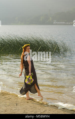 Junge Frau mit gelben Blumen in der Hand am Ufer des Sees, langen schwarzen Kleid, mit Ihrem rechten Fuß zum Wasser erweitert. Stockfoto