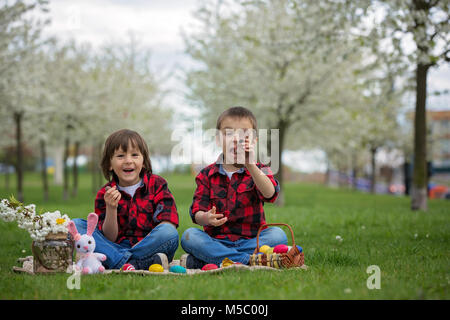 Zwei Kinder, junge Brüder, Schokoladenhasen Essen und Spaß mit Ostereier im Park, schön blühenden Frühlingsgarten Stockfoto