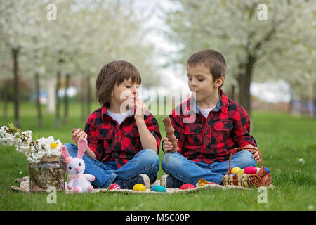 Zwei Kinder, junge Brüder, Schokoladenhasen Essen und Spaß mit Ostereier im Park, schön blühenden Frühlingsgarten Stockfoto