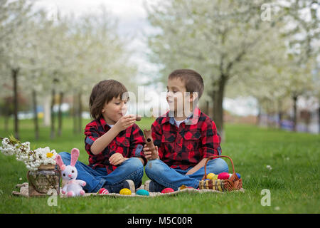 Zwei Kinder, junge Brüder, Schokoladenhasen Essen und Spaß mit Ostereier im Park, schön blühenden Frühlingsgarten Stockfoto