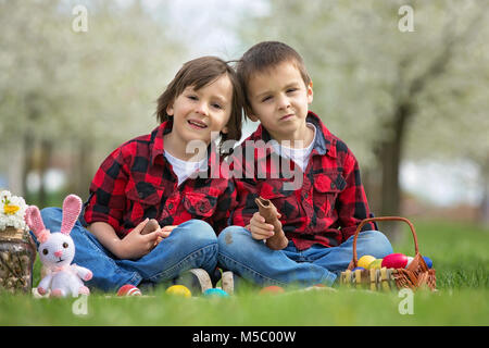 Zwei Kinder, junge Brüder, Schokoladenhasen Essen und Spaß mit Ostereier im Park, schön blühenden Frühlingsgarten Stockfoto