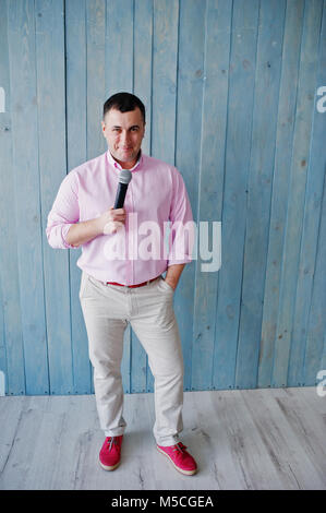 Schöner Mann in rosa Shirt mit Mikrofon vor blauem Holz Wand Hintergrund im Studio. Toastmaster und Showman. Stockfoto