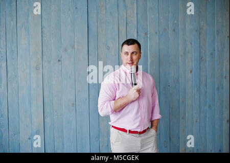 Schöner Mann in rosa Shirt mit Mikrofon vor blauem Holz Wand Hintergrund im Studio. Toastmaster und Showman. Stockfoto