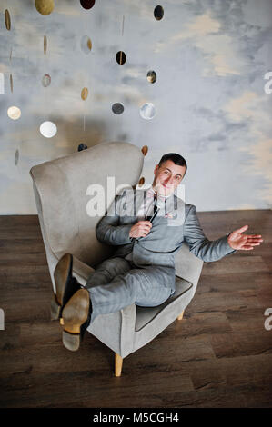 Schöner Mann im grauen Anzug mit Mikrofon sitzen auf Stuhl Hintergrund Studio. Toastmaster und Showman. Stockfoto