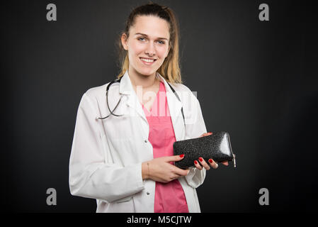 Weibliche junge Doktor holding Wallet und lächelnd auf schwarzen Hintergrund mit Copyspace Werbefläche Stockfoto