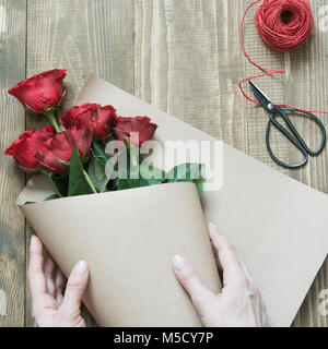 Florist eine rote Rosen Blumenstrauß, Umwickeln mit Kraftpapier auf einem Holztisch. Blick von oben. Im rustikalen Stil. Stockfoto