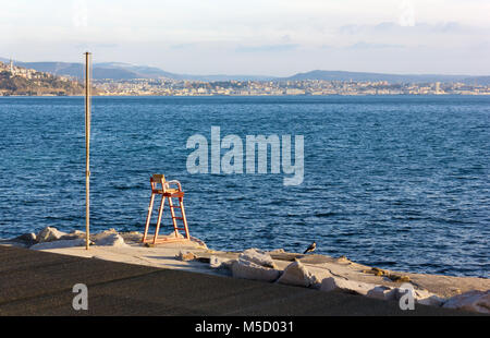 Winter Blick auf ein Rettungsschwimmer Stuhl an der Küste von Barcola Bezirk in Triest, mit der Skyline der Stadt im Hintergrund Stockfoto