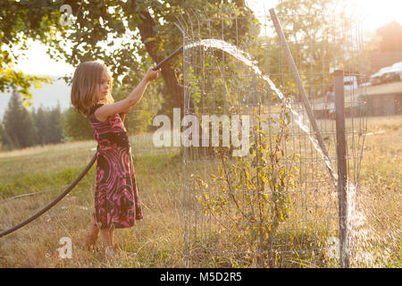 Mädchen im Kleid Bewässerung Baum mit Schlauch im sonnigen Sommer Yard Stockfoto