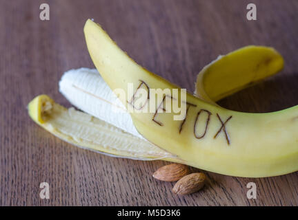 Nahaufnahme der gelben Banane mit geschriebenen Wort Detox und zwei Mandeln liegen auf hölzernen Tisch Hintergrund Stockfoto